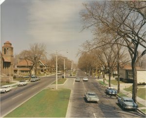 Elevated view of North Sherman Boulevard from North Avenue in daylight. The avenue is divided by a central grass median. Some cars traverse the avenue, and some are parked on the sides. Tall trees grow on the road verges. Sidewalks and buildings in the vicinity are visible.