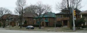 Panoramic view of multiple-story dwellings and other buildings along North Sherman Boulevard. Several leafless tall trees grow on the road verge along the avenue. A car is parked in front of the building in the middle.