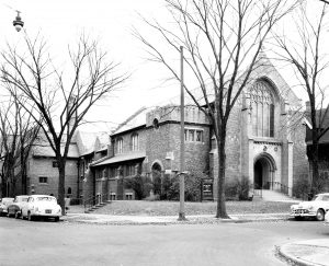 Grayscale long shot of Plymouth Church UCC building sitting on the street corner. The image shows the church facade and side. The facade is on the right, featuring an arched entrance and a large arched window above. The side is on the left, featuring a smaller arched entrance and several windows. An intersection and cars parked on the street's side are visible in the foreground. Leafless tall trees grow on the road verge.