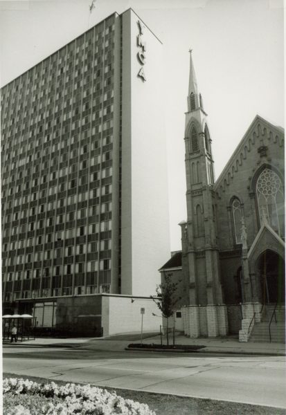 Sepia-colored photograph of the YMCA tower on the left. The facade faces slightly to the left. The building's side has a vertical "YMCA" sign on its top. The Calvary Presbyterian Church is on the right. A bus stop appears in front of the YMCA tower. A street is visible in the foreground.