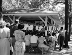 People sit on wooden benches surrounding a pavilion's edge while playing bingo. Two people stand inside the pavilion, one holding a microphone. A sign that reads "play bingo" hangs fromthe pavilion's ceiling. Trees shade the structure's roof and the surrounding area. Some people stand in the left foreground, watching the game from a distance.