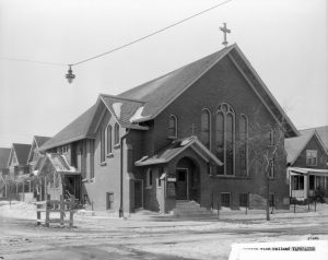 The frontage of the Christ Polish Baptist Church located on a streetcorner adjacent to residential buildings. The church has a broad gabled roof and a cross erected on top of it. A gabled entry porch and tall, narrow arched windows are some of the main features of the building's exterior. Snow surrounds the church and covers some parts of the street. A deciduous tree, bare of foliage, stands near the sidewalks in front of the property.