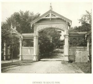 Sepia-colored long shot of the open Schlitz Park entrance gate on unpaved road. It consists of two roofed gates. A smaller one on the left and the larger one for a driveway on the right. On the top front of the large gate is inscribed "Schlitz Park." The whole structure is predominantly made of wood.