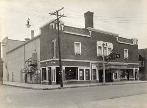 Long shot of Riviera Theater by a street corner in sepia. The facade faces slightly to the right. The theater's front sign sits atop the marquee above the main entrance. The two-story building features a chimney on top of the roof surrounded by crisscrossing overhead wires. A combination of columns and rectangular and display windows compose the ground floor. A vertical sign that reads "Soda" is on the building's left upper side. Roadways in the foreground are empty.