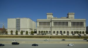 Long shot of Milwaukee County Courthouse standing in the distance against a clear blue sky. To the left of the courthouse is a large complex that houses the Milwaukee County Jail and the county Safety Building. Cars pass through a highway in the foreground.