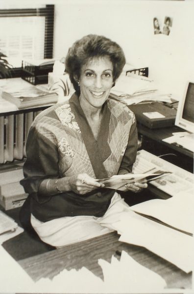 A sepia-colored medium shot of Ellen Bravo from the thighs up smiling in a casual top while holding papers or photographs with both hands. She is sitting next to two wooden office desks surrounding her. Between 2 o'clock and 4 o'clock, on one of the desks, are a computer monitor and a keyboard. Close to it are two small photographs hung on the wall. Some documents are piled on the desk in front of Bravo. Some others appear in a less neat pile behind her. A window with open blinds attached to the wall between 10 and 11 o'clock.