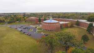 Bird's eye view of Sacred Heart Seminary and School of Theology campus surrounded by green landscape. The image's center shows the iconic round Sacred Heart Chapel that features a light blue-colored roof with a crown and a cross atop. Behind the chapel, from the image's center to the right, is a grand multi-story building whose shape resembles a semicircle. Trees grow in the background as far as the eye can see. Several cars are parked in front of the chapel. Expansive green lawns and trees embellish the front yard.