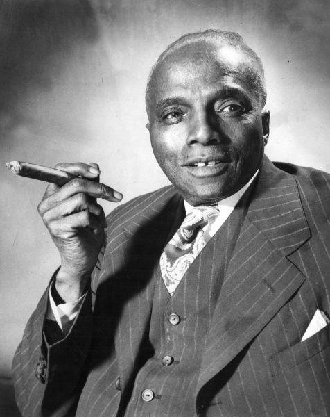 Headshot of J. Anthony Josey from the elbows up smiling in a vertical striped vest and notched lapel suit, with a paisley cravat. Josey's right hand's index and middle fingers elegantly clamp a cigar while his face and eyes look to his left.