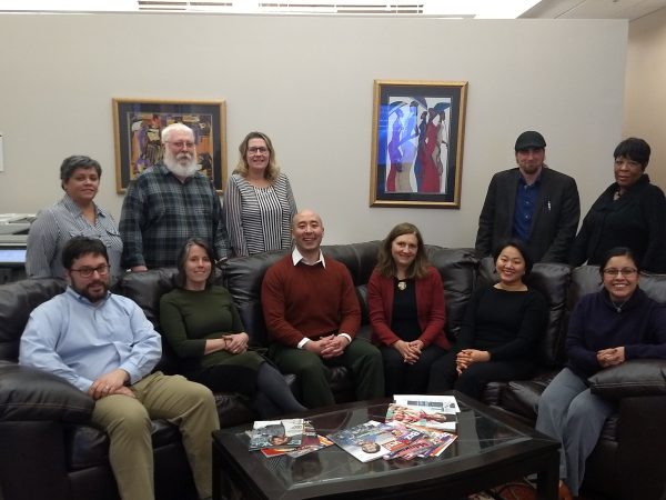 Group photo of the Legal Aid Society of Milwaukee members in an indoor space at their downtown office. Six people sit on a black L-shaped couch. Three members stand behind the couch on the left and two on the right. They smile while making direct eye contact with the camera lens. Two paintings hang on the white wall behind.