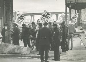 Sepia-colored full shot of a group of people in warm clothes walking to the left. Some carry protest signs that read "On Strike, Local 248 Amalgamated Meat Cutters AFL-CIO." Parked cars appear in the background. The bottom part of a freeway is visible in the far distance.