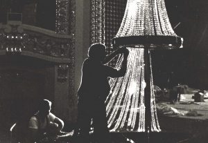 Silhouette of two people working on a large crystal chandelier in the Pabst Theater. The image is in grayscale. One of the people stands at the image's center with hands touching the sparkling crystals. The other one appears on the left and seems to kneel while working on the chandelier's bottom part. Two arched openings are visible in the background. Each is embellished by various small lights highlighting the arched structure of the openings.