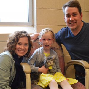 A woman in a green jacket and a man in a dark blue T-shirt flank a young girl in a yellow dress and grey cardigan who sits on a chair while holding Buffy, the kitten. They are Stacey, Sally, and Jim. They smile as looking at the camera lens. A window and wall are in the background.