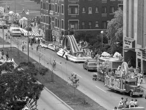 High-angle shot of Wisconsin Avenue during the 1983 inaugural City of Festivals Parade shows a line of floats stretching down the road. The crowd on the sidewalks gazes at the floats decorated in various themes, shapes and sizes.