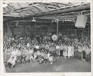 Grayscale elevated view of dozens of Regal Ware employees posing for this photograph inside the Kewaskum factory. People stand and sit on the floor and chairs appear here and there, filling the space. Many make eye contact with the camera lens. The steel ceiling structure is visible.