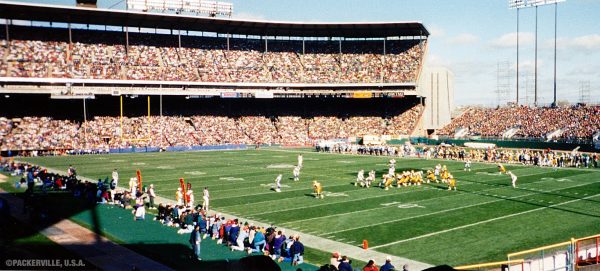 Wide shot of two football teams playing at the County Stadium. Lines of photographers kneel by the green football field while aiming their camera at the players, who are in action. Sun rays hit the field and the packed grandstand.