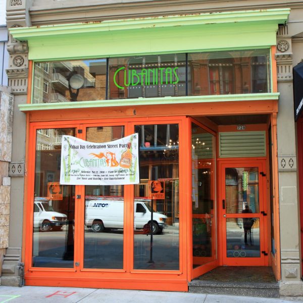 Main entrance of Cubanitas restaurant features soft green and orange exterior walls. A banner advertising the Cuban Day Celebration Street Party hangs on its glass wall. White vans parked on the street are reflected in the restaurant's plate glass windows.