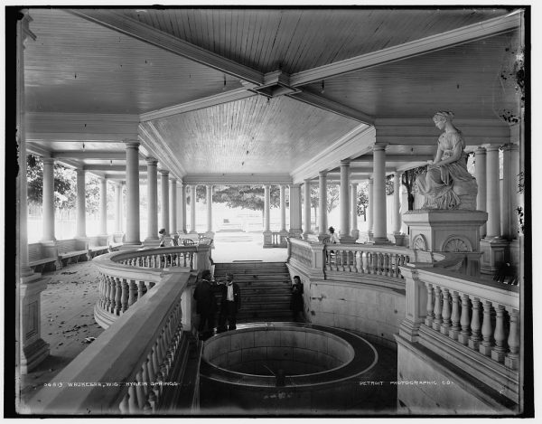 Grayscale photograph of visitors to Hygeia Springs. The pavilions' interior, with the spring well in the center foreground, is showcased in this image. The stairway down to the well is in the center back. Some people stand around the stairs. Wall and balustrades surround the stairway and the area around the well. The Hygeia sculpture sits on the upper floor on the right of the well. The upper story's floor area is visible on the left. The ceiling is visible. Rows of columns and outdoor views can be seen in the background.