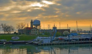 Panoramic view of Port of Kenosha and Lake Michigan against a glowing orange sky. Rows of boats are visible from the center to the right foreground. The iconic Southport lighthouse tower and the red roof lighthouse keeper's residence building appear in the distance. Appearing next to the lighthouse tower is a large water tank decorated with icons of the lake, trees, and sailboats.