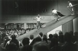 Three men in casual clothes demonstrate skiing on a man-made sloping terrain installed inside the mall. They hold ski poles and wear skis on their feet. Onlooker crowds around them are gazing up at the demonstration.