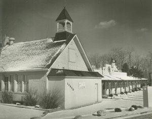Grayscale long shot of the Brookfield building exterior view in sepia. The structure on the left has a bell tower atop its roof. A one-story structure appears on the right background featuring regularly spaced wooden porch posts. Snow covers the ground and part of the roofs. Trees bare of foliage are visible in far right background.
