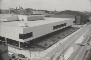 Grayscale elevated view of the corner of MECCA building. The building is clad in vertical white panels punctuated by banks of black windows at the corners and center. A mechanical penthouse-like structure sits atop its flat roof. Cars enter its covered driveway. The building now known as the UW-Milwaukee Panther Arena is located next to MECCA in the right background, on the next block.