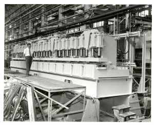 Long shot of Nordberg Manufacturing Company interior in grayscale. The image shows a man in a dress shirt, tie, and trousers standing on the left on a wooden scaffolding while looking at a large molding press spanning next to him. Multiple pipes appears in the upper background, extending from left to right.