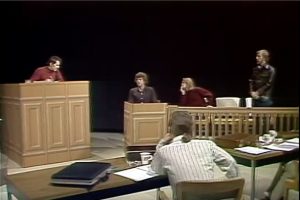 Long shot of Theater X actors performing on stage with a black backdrop. The stage set and props illustrate a courtroom. Four people perform in the image's background. Willem Dafoe in the red sweater is third from the left. One person sits behind a long desk in the foreground.