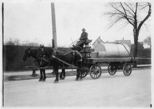 Grayscale long shot of a man sitting atop a horse-drawn wagon carrying a metal tank in an empty street. The two horses that pull the wagon face to the left. The man holds the reins while making eye contact with the camera lens. A long wooden fence appears in the background. A building is visible on the far right back.