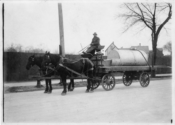 A man sits atop a horse-drawn wagon carrying a metal tank. The wagon is labelled as belonging to the Heil Company, located in the Layton Park neighborhood.