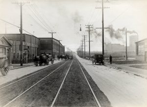 Taken outside the Globe Seamless Tubes Company around 1922, this photograph showcases both horse-drawn wagons and automobiles stationed on either side of electric streetcar tracks.