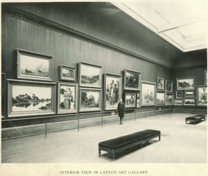 Sepia-colored long shot of Layton Art Gallery interior showcases framed paintings hanging on the walls. One of the room's corners appears on the right. Stanchion posts run from left to right to protect the works of art. Someone in formal attire stands at the image center with their right hand holding on to the stanchion. Two gallery benches sit on the floor—one in the center foreground, one in the far right. The room's ceiling is visible. Text at the bottom of the photo reads "Interior View in Layton Art Gallery."