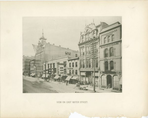 Wide shot of East Water Street showing a rows of commercial buildings ranging from two to four stories. Store signs are displayed in front of each business place. A group of people stands on the porch of the "J.C. Iversen & Co." building. Two horse-drawn vehicles stand by the sidewalk. Two large utility poles also stand in the street next to the sidewalk.