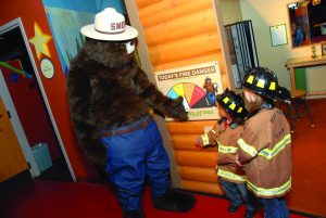 A person in a "Smokey the Bear" points to a forest fire-related learning board while interacting with two children dressed up in hotshot crew uniforms. The US wildfire prevention mascot wears blue jeans, a brown belt, and a round hat bearing the name "Smokey." They are standing in a colorful exhibit arena.