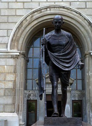Full shot of the bronze statue of Mahatma Gandhi in his iconic cloth and glasses holding a walking stick. Behind the sculpture is one of the entrances into the Milwaukee County Courthouse.