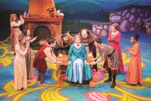 High-angle shot of actors performing "Ella Enchanted" in colorful costumes on a colorful indoor stage. In the center, an actress in a blue dress sits on a table with her mouth open, while four actors surround her making the same expression. Three other people on the left and three on the right clap their hands while looking at the actress in the center.