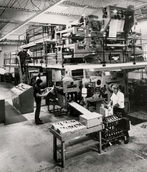 Grayscale elevated shot of three men working inside the Quad/Graphics Pewaukee plant. A set of large machines sits on two-level of platforms in the background. Steel structure in the ceiling is visible. A man in the left background walks down the stair attached to the platform. Two other employees stand in the foreground in the area below the machine. Both check on the papers they have printed. The man in a headset on the farthest right stands at the end of the machine that releases printed products. His hands are on top of a stack of the products.