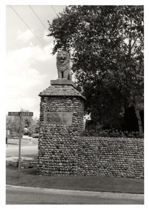 Image of the Lion Gate that features a lion statue placed on top of stone walls. Written on the sculpture's base is the name of one of its creators, which is "Fred Usinger." On the stone wall, several feet beneath the statue is inscribed "Country Life in Donges Bay 1890 – 1911.” Next to the gate is a street name sign that reads "E Fairy Chasm Rd." Lush trees growing behind the stone walls appear in the right portion of this image.