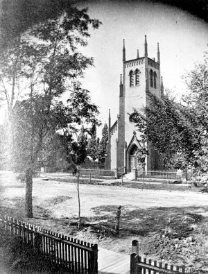 Grayscale long shot of North Presbyterian Church facade sitting across a street in the background. Its central tower and entrance are visible in the distance. A fence surrounds the front yard. Trees grow next to the church and on the road verge in the image's foreground. A fence can be seen in the foreground.