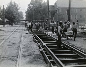 Grayscale long shot of a group of men working to lay streetcar tracks along N. 8th Street. They work on the tracks that stretch from the background to the right foreground. Other tracks span to the left foreground. Heavy equipment can be seen in the street side on the center back. Tall trees grow on either side of the street. A building is seen in the far right in the background.