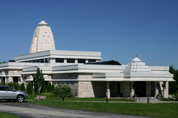 Exterior view of Hindu Temple of Wisconsin in Pewaukee against the blue sky. The outer walls of the temple are dominated by white and cream colors with a tower in the background. The building is surrounded by a green lawn and plants.