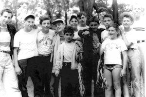 Grayscale group photo of boys standing close to each other in an outdoor space. The kids smile in their summer clothes. Some put their arms around others' shoulders, showing a friendly gesture. Two in the front hold up fish on their fishing lines.