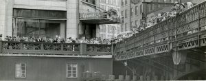 Long shot of crowds gathering on the railings of a pedestrian area that is located on the river bank and the Wisconsin Avenue bridge. They gaze down at Gertie and her ducklings that sit on the wooden piling below. The Gimbels entrance sign and a large display window are visible on the left background.