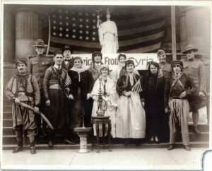 Reproduction of a sepia-colored group photo of thirteen Milwaukee Syrians posing on stairs. Some hold the USA stick flags. A large American banner appears in the background behind a statue. This photo is slightly torn on the lower right side.