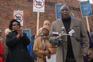 Medium full shot of Rev. Willie Brisco in a suit standing on the right while giving a speech with his right hand holding a microphone. His body faces directionally to the camera. Two other mics owned by broadcasting companies are set in front of Rev. Brisco. A crowd of people stands behind him. Some hold protest signs. A woman in glasses on the farthest left claps her hands