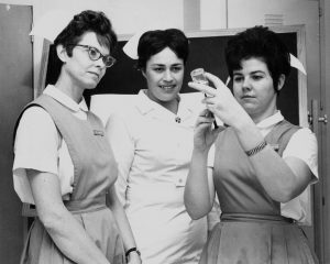 Grayscale medium shot of three people standing in uniforms. The woman in the center wears a different uniform than the two who flank her. The nursing student on the right draws liquid from a vial into a syringe while the rest of them pay attention to the process.