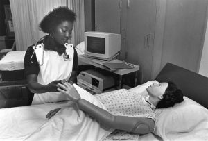 Grayscale portrait of Carol Alston standing and a mannequin lying on a patient bed in a patient room. Alston holds the mannequin's left hand while wearing a stethoscope around her neck. Behind her are another bed and a curtain partitioning the room. A computer sits on a table next to Alston.