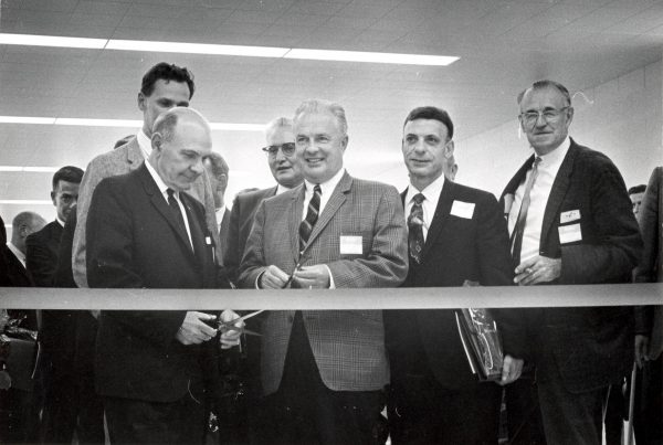 John Doyne stands among men in suits and ties, smiling as he holds a pair of scissors for a ribbon-cutting ceremony. Another group of men appears in the background. A thick ribbon stretches horizontally in front of them.