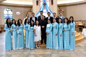 Group photo of Father Joseph Quang and several choir members posing in two rows inside St. Martin of Tours church. Father Quang, in black-colored attire, stands in the center, between the first and second rows. Female choir members in light-blue and white dresses stand in the front. Six male members stand in the back. They are in white shirts and light-blue ties. All men are in suits except the one on the far left. A wooden cross is attached to the wall in the background. Sunshine goes through a mosaic window that appears on the left background.
