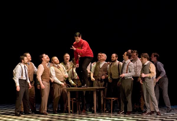A group of male actors gathers in a semi-circle surrounding a man standing on a table in a bright red sweater. Members of the crowd show different facial expressions in their vintage clothes. Some gaze up at the actor in red. The background is pitch black, and the floors are in a checkerboard design.