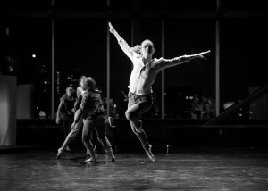Black and white long shot of dancers in action. The center of this image shows one of the dancers in a jumping position with both hands stretched up looking like wings. Three others appear in the left background.